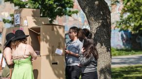 Trois élèves présentent une structure composée de boîtes de carton à deux femmes dans un parc.