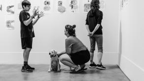 Un élève discute avec une femme qui flatte un chien. Une autre femme regarde les dessins exposés. Crédit @ Katia Konioukhova.